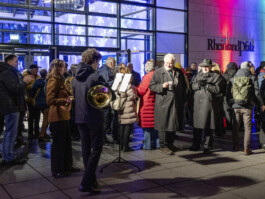 Gruppe von Menschen vor dem Eingang der Vertretung der Rheinland-Pfälzischen Botschaft in Berlin bei einer Weihnachtsfeier, einige hören einer kleinen Blaskapelle zu.